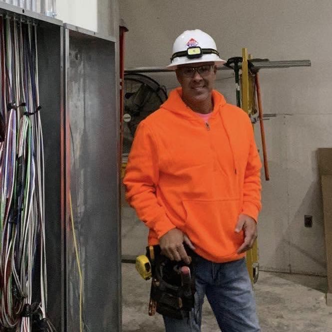 Tony in a hard hat and tool belt beside an electrical panel